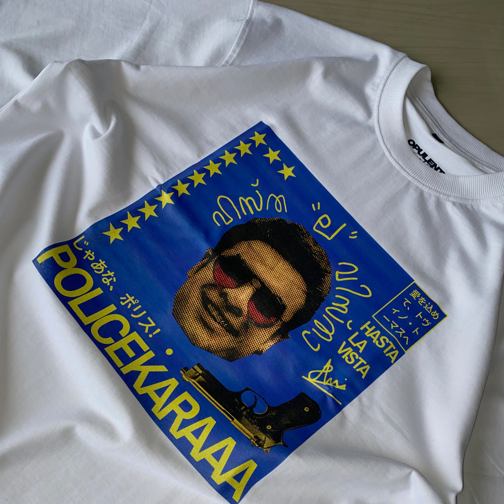 The Tovino Policekaraa is a white, oversized 220 GSM sweatshirt with a colorful graphic of a man's face in sunglasses, yellow stars, various languages, a handgun, and bold “POLICEYKARAAA” and “HASTA LA VISTA” text on a blue background.