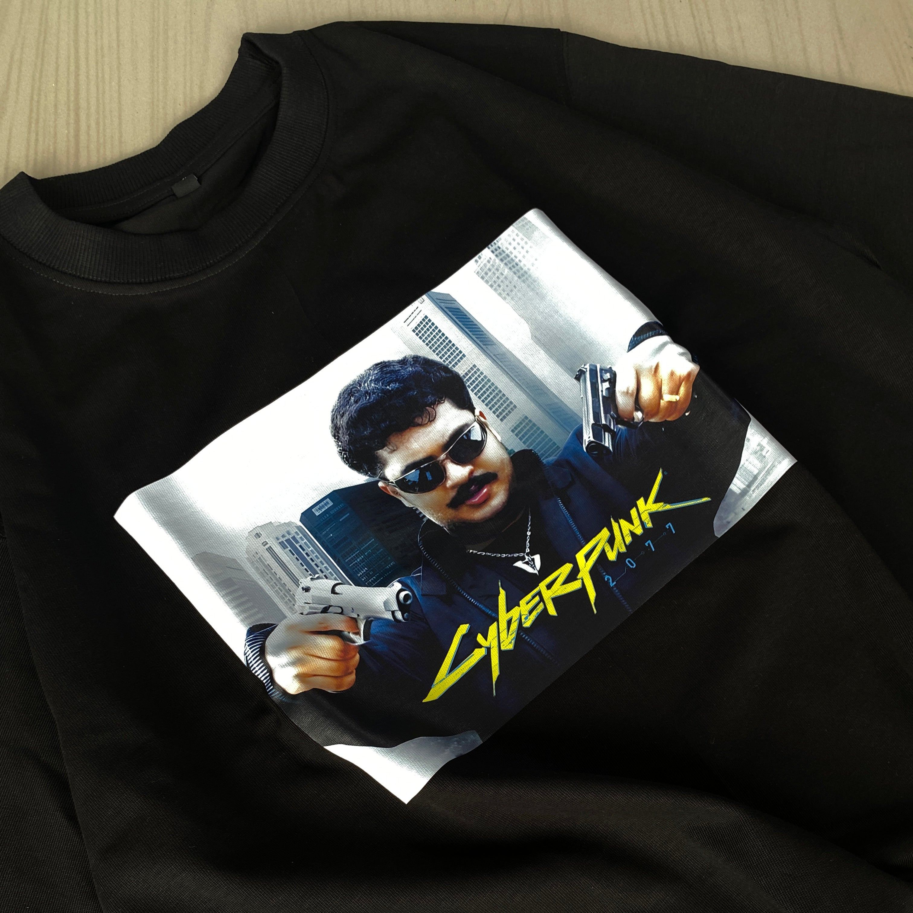 The Cyberpunk VINU is an oversized black t-shirt made from heavy weight 220 GSM fabric, showcasing a man with sunglasses and two guns against city buildings, with yellow Cyberpunk 2077 text below.