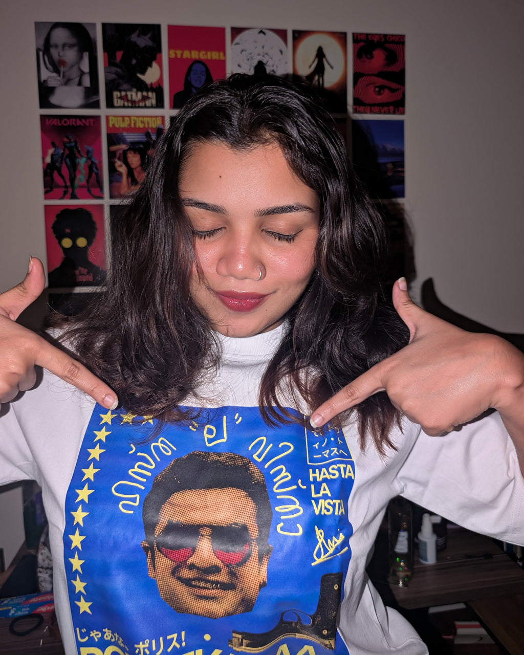 A woman with long dark hair smiles, eyes closed, and points at her oversized white "Tovino Policekaraa" T-shirt featuring a colorful graphic of a man's face in sunglasses and stylized text; posters hang on the wall behind her.