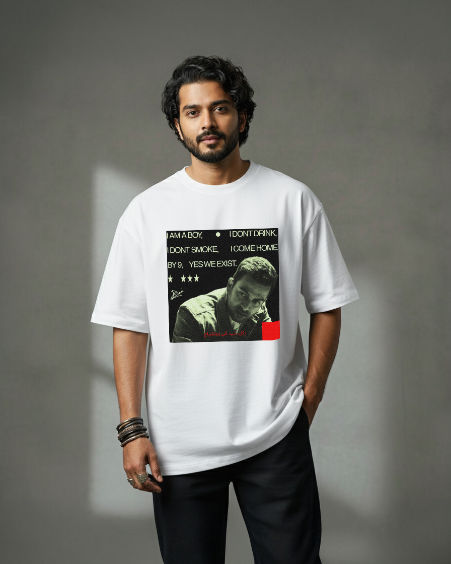 A man with dark, wavy hair and a beard stands against a gray backdrop, wearing the oversized GoodBoy Prithvi white T-shirt featuring a black-and-white graphic and text, paired with black pants and bracelets on his left wrist.